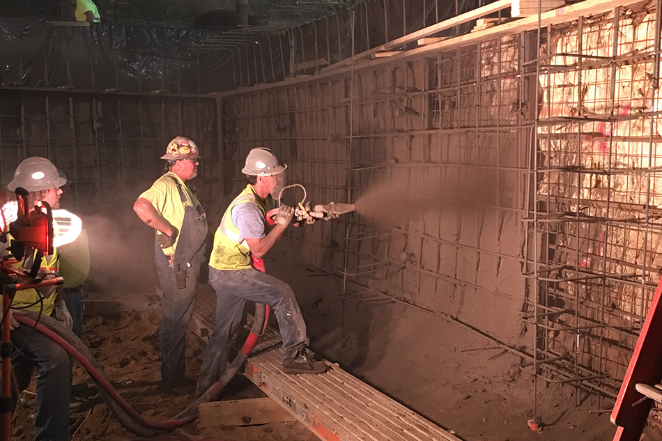 Three construction workers in a dark building spray concrete to build a wall, illuminated by work lights