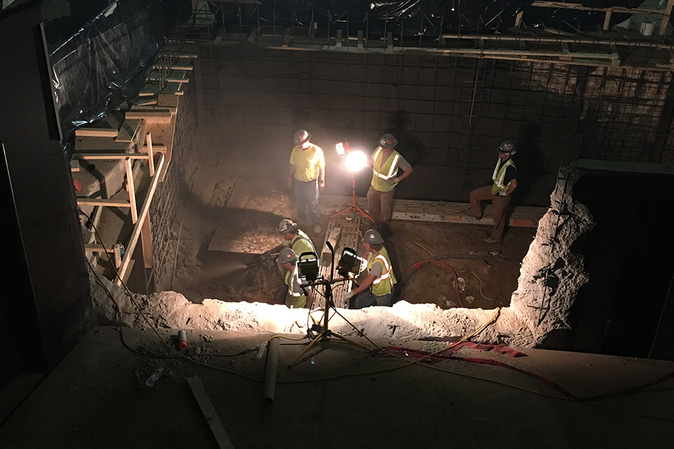 Construction workers in a dark building spray concrete to build a wall, illuminated by work lights
